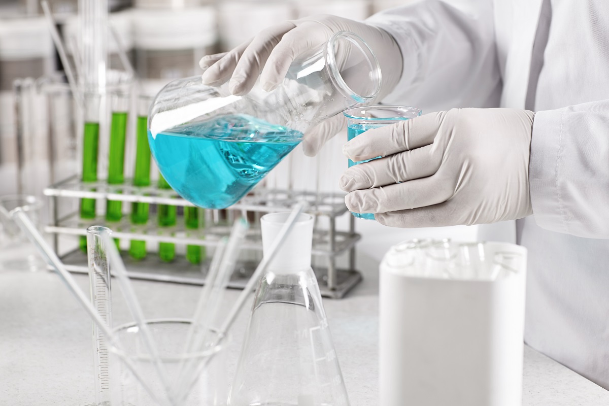 Young clinical worker dressed in white gown and gloves holding glass beakers with blue liquid making experiment in laboratory. Scientific laboratory research, laboratory beakers in hadns of scientist